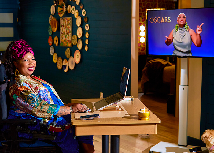 Andraéa LaVant, a pecan-brown Black woman in a bright blue jumper, patchwork kimono, and fuchsia headwrap, smiles wide at her desk with one hand on a mouse beside an open laptop. Behind her, a teal wall features circular plate art and a painting. A TV nearby shows a Black Deaf woman interpreter signing, with the Oscars logo onscreen. Goji’s head peeks in from the corner.