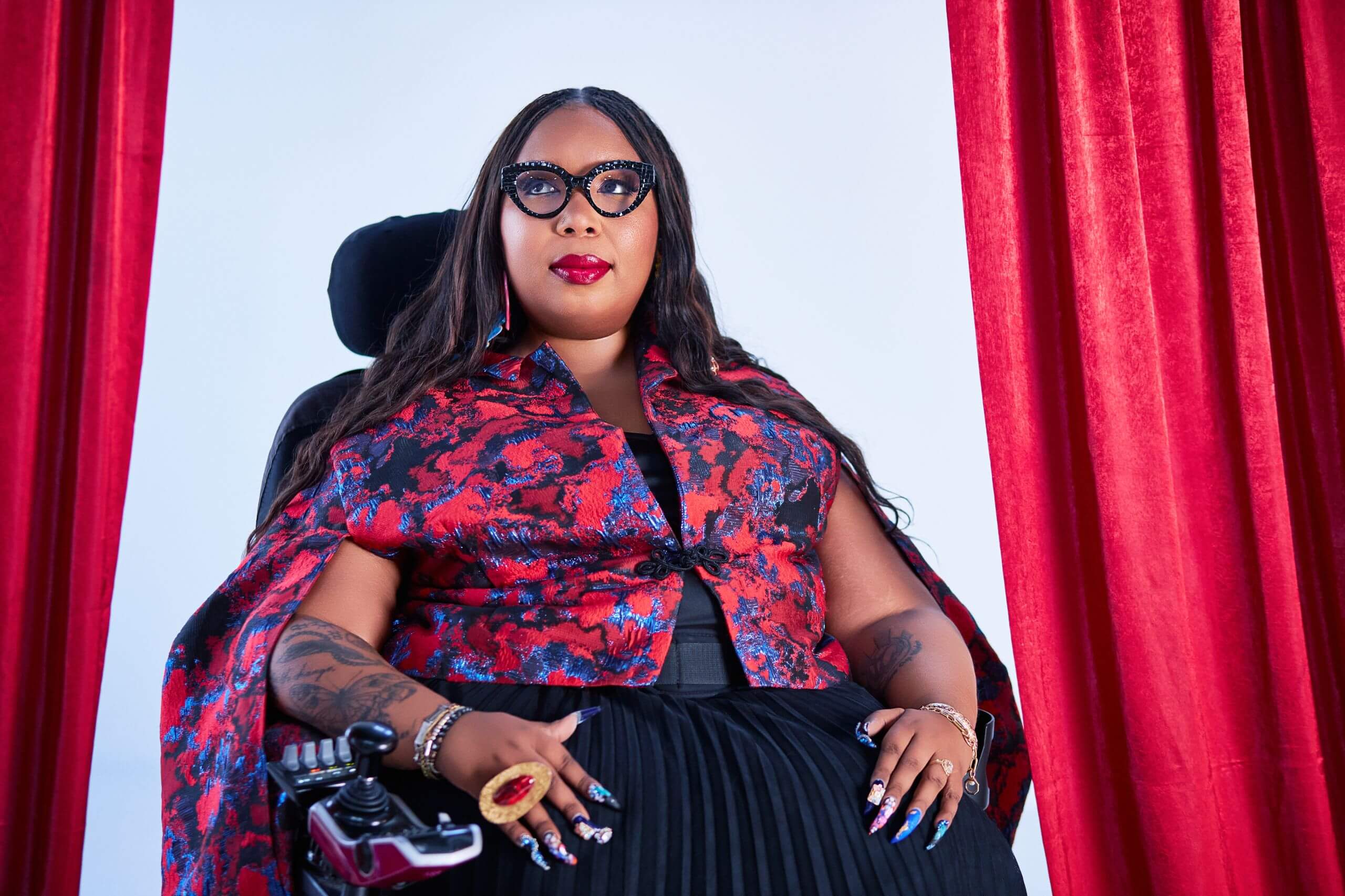 Andraéa LaVant, a pecan-brown Black woman with dark brown mermaid braids, sits in her power chair wearing black cat-eye glasses, a black chiffon dress, and a red and blue jacquard capelet. Her glossy red lips, extravagant nails, and bold jewelry shine as she gazes upward, framed by open red velvet curtains.