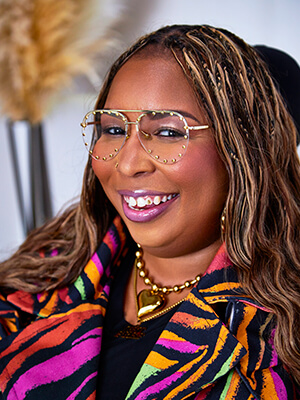 Close-up of Andraéa LaVant, a pecan-brown Black woman with two-tone mermaid braids, smiling wide in profile with a visible silver tooth gem. She wears studded aviator glasses, pink lip gloss, and a beaded gold heart necklace over a neon zebra trench, with a hint of black dress showing. The softly blurred backdrop includes tan feathers and a white wall.