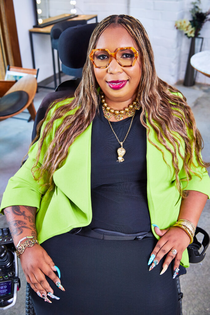 Andraéa LaVant, wearing ombré-orange hexagon glasses and fuchsia gloss, looks up at the camera with a slight smile. Her two-tone mermaid braids frame a chartreuse blazer and gold jewelry. Behind her, a lit vanity mirror sits on a desk.