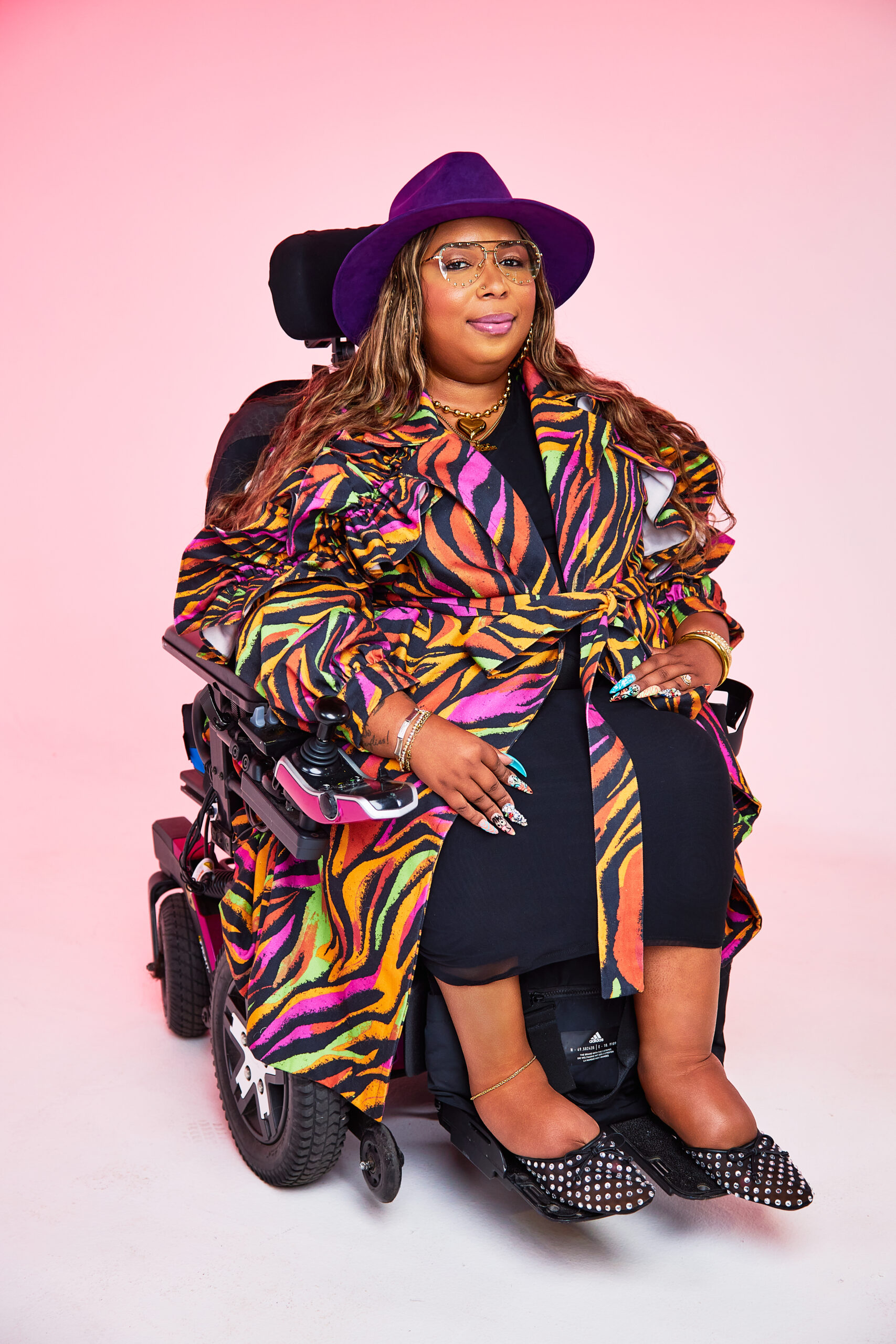 Andraéa LaVant, a pecan-brown Black woman, smiles slightly with closed lips, seated in her fuchsia and white power chair wearing a royal purple fedora and studded aviator glasses. A neon zebra trench, jeweled nails, and rhinestone slippers stand out against a pinky-peach ombré backdrop.