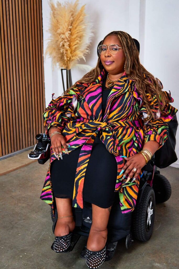 Andraéa LaVant, a pecan-brown Black woman, smirks in pink lipstick and studded aviator glasses, side-eyeing from her fuchsia  and white power chair. A neon zebra trench, jeweled nails, and rhinestone slippers stand out against a soft, textured backdrop.