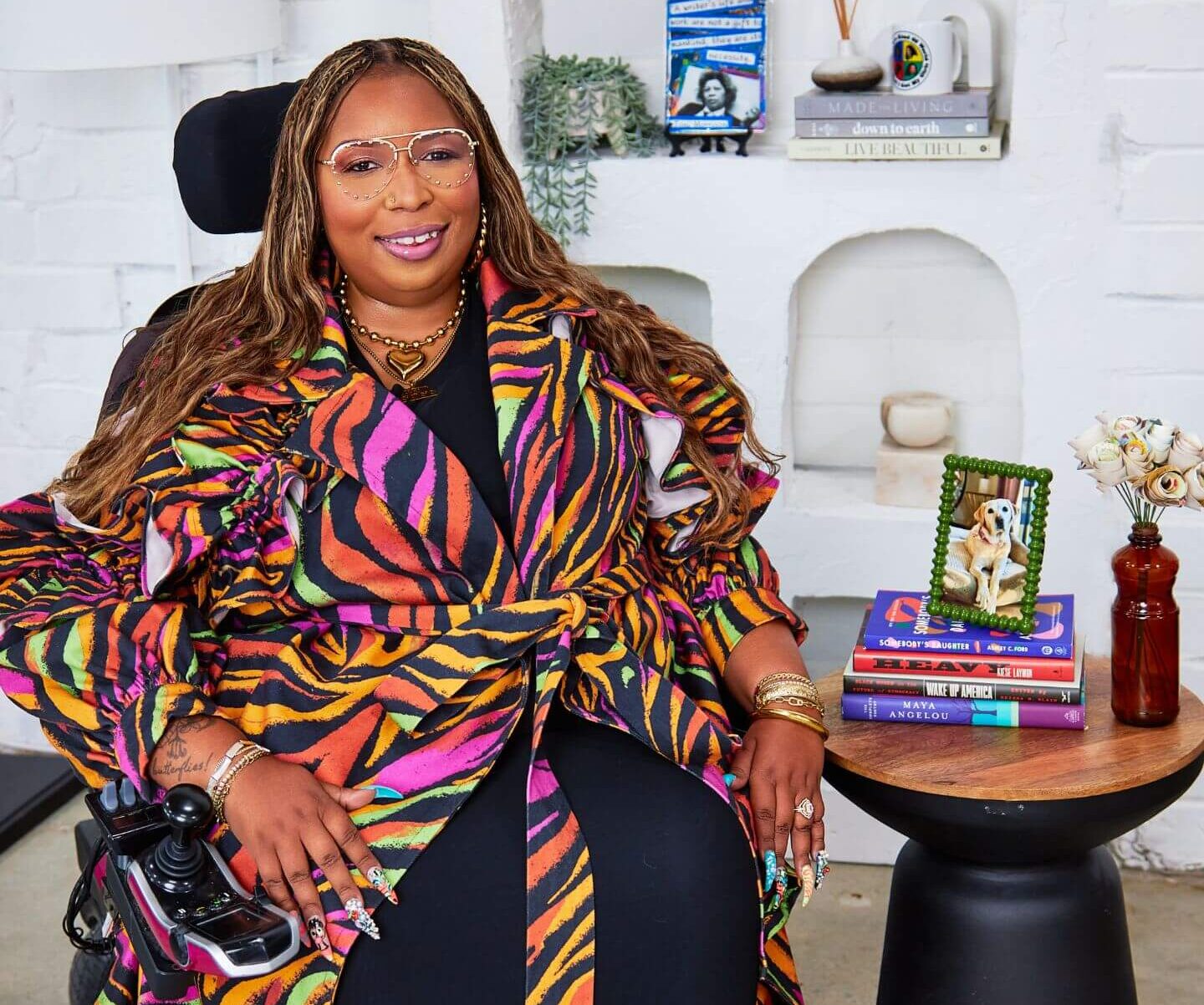 Andraéa LaVant, a pecan-brown Black woman in a neon zebra trench and studded aviators, sits in her power chair beside a round table with colorful books, paper flowers, and a framed photo of her dog, Goji. Behind her, arched inlets in a white stone wall hold books and a plant, including a visible Toni Morrison journal.
