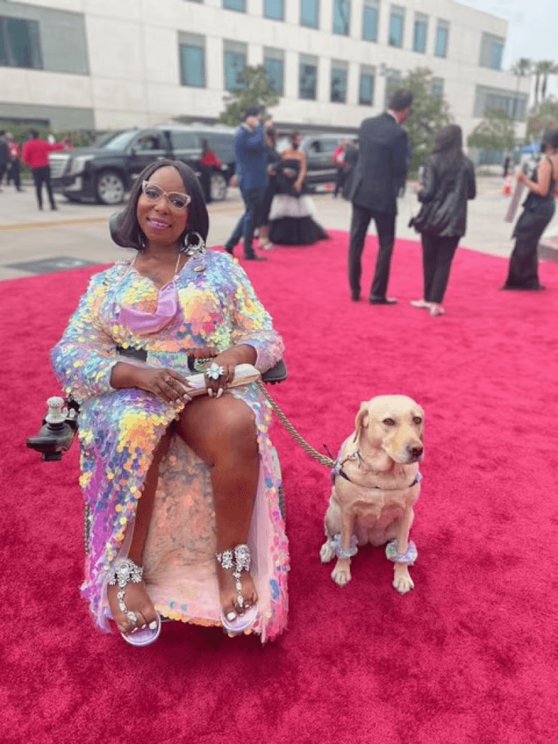 Andraéa LaVant sits confidently in her wheelchair on a red carpet, smiling in a pastel iridescent sequin gown and jeweled sandals. Her hair is sleek and shoulder-length, and she wears bold cat-eye glasses and statement earrings. Beside her sits Goji, her yellow Labrador service dog, adorned with lavender tulle accessories on her front paws.