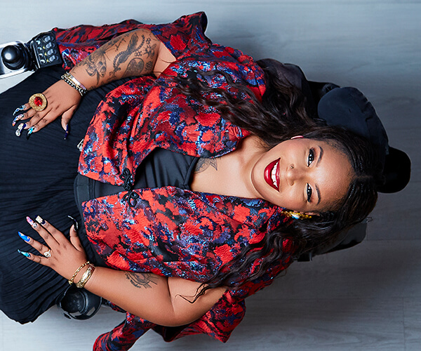 Andraéa LaVant, a pecan-brown Black woman with dark brown mermaid braids, smiles wide as she leans all the way back in her power chair, looking up at the camera. Seen from above, knee-up, she wears black cat-eye glasses, a black chiffon dress, and a red and blue jacquard capelet, with bold jewelry and glossy red lips.